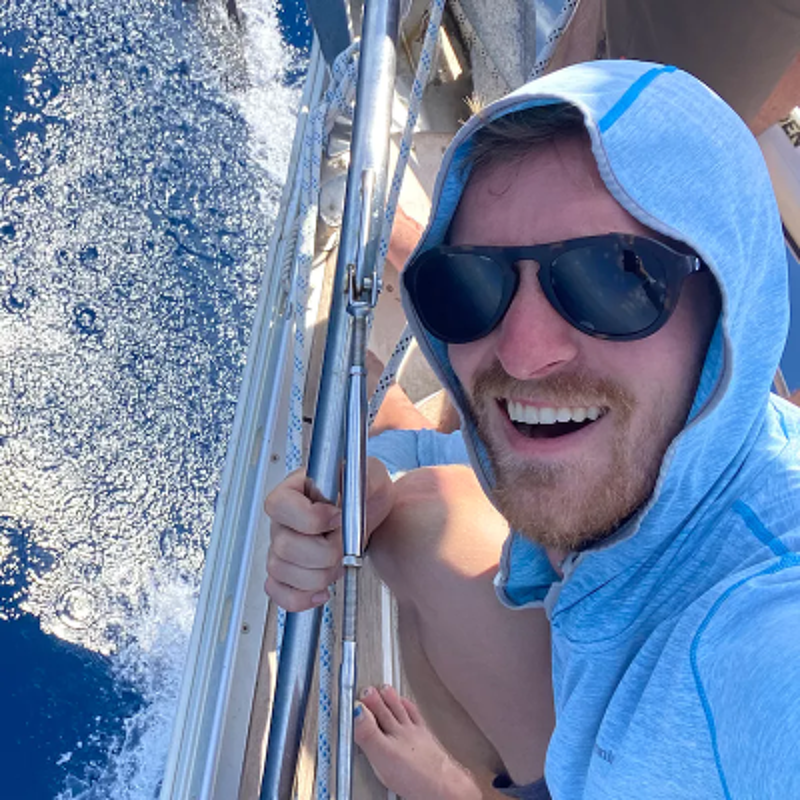 The image shows a man on a sailboat, smiling at the camera. He is wearing sunglasses and a hooded sweatshirt. The background shows the ocean and the boat's rigging. The man appears to be enjoying himself on a sunny day. The water is choppy, and there is a hint of rain in the distance.